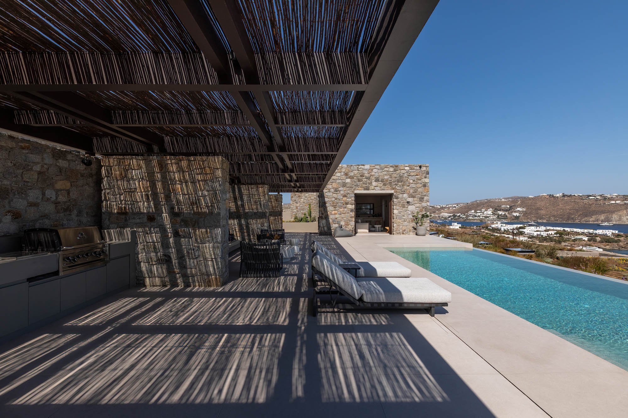 Sun loungers under bamboo pergola, infinity pool and Mykonos bay beyond