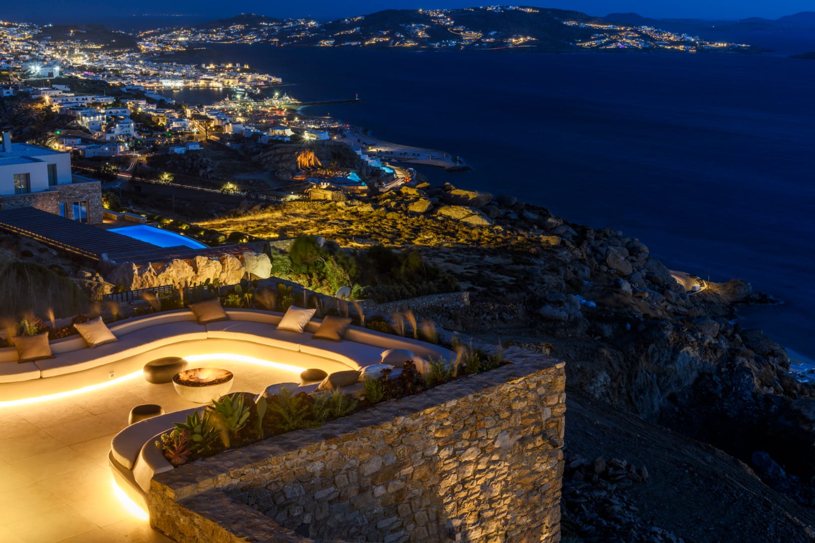Stone fire pit lounge at night, the lights of Mykonos town below