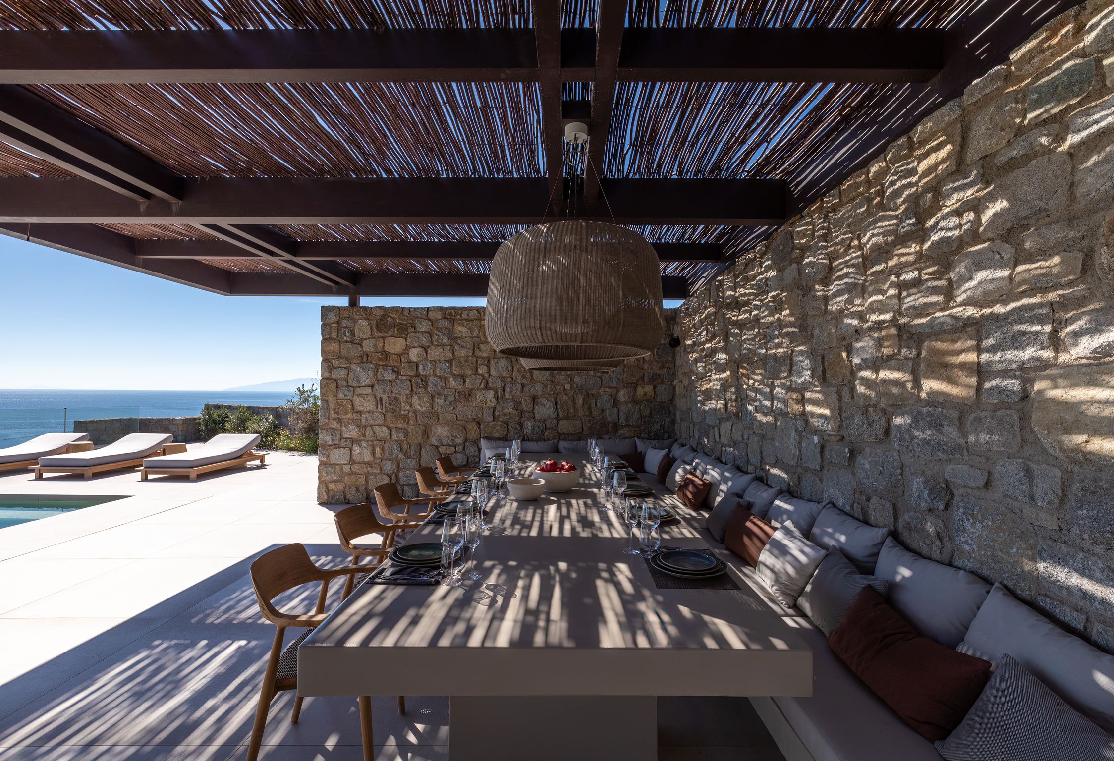 Long outdoor dining table beneath a bamboo pergola and stone wall